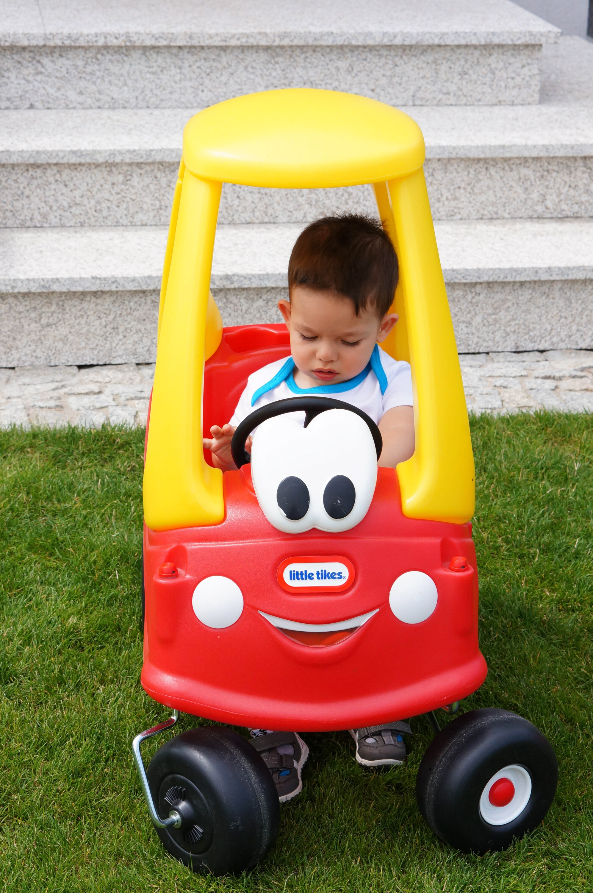 Small boy sitting in a Little Tikes plastic wheeled Cozy Coupe cart having a yellow superstructure, steering wheel, googly eyes and a red body with white circles simulating headlights, the Little Tykes logo where a nose might be below the eyes, and a smiling mouth under the logo.