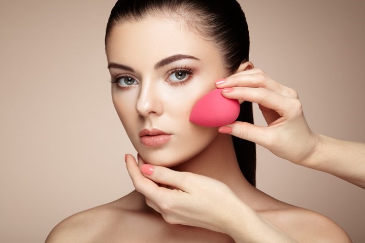 Photo of the face of a model having makeup applied by female hands holding pink pear-shaped Beautyblender sponge.