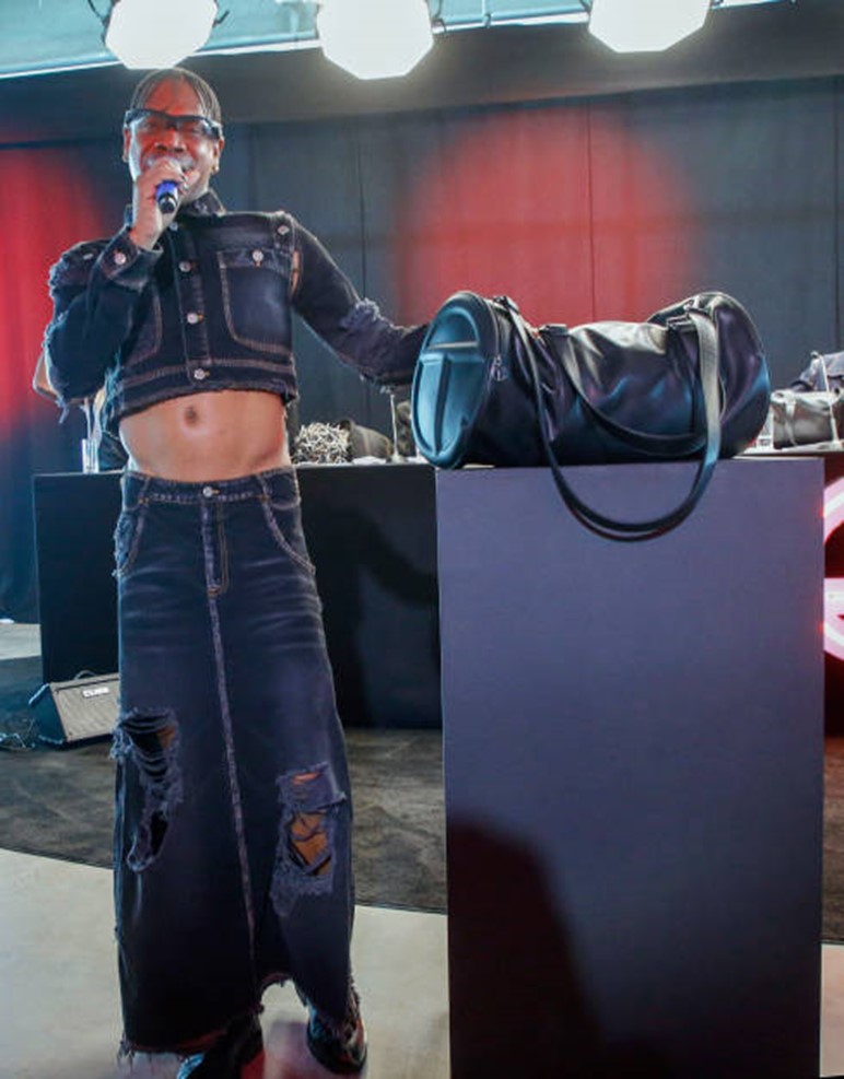Photo of Telfar Clemens speaking into a wireless microphone in his right hand, his left hand on top of a black duffelbag sitting on a stand, and wearing a short dark blue denim jacket with a bare midriff and loose-fitting dark blue denim pants having patches on the legs.