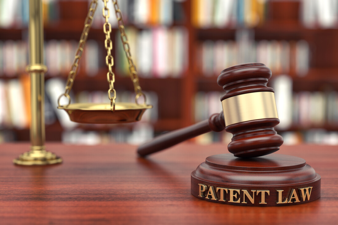 Gavel resting on a pedestal with Patent Law on the side of its base on a table next to one basket of a scale of justice in front of a wall of bookshelves